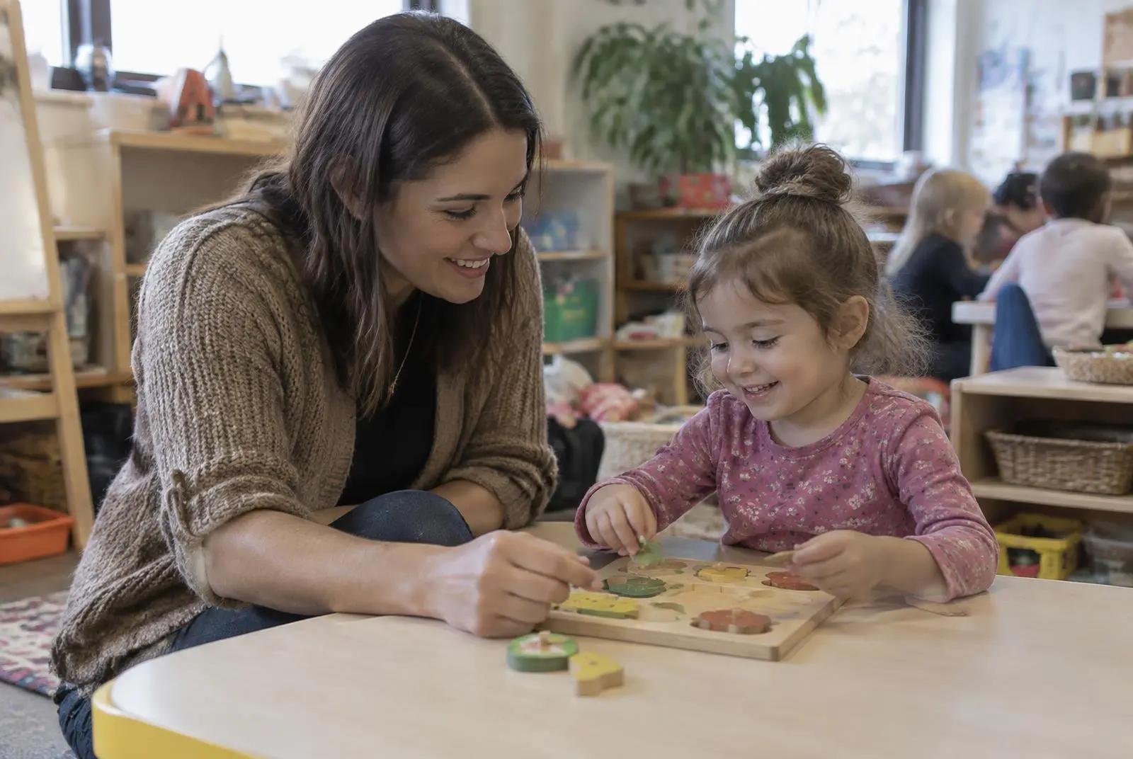 A smiling teacher helps a preschooler with a wooden puzzle